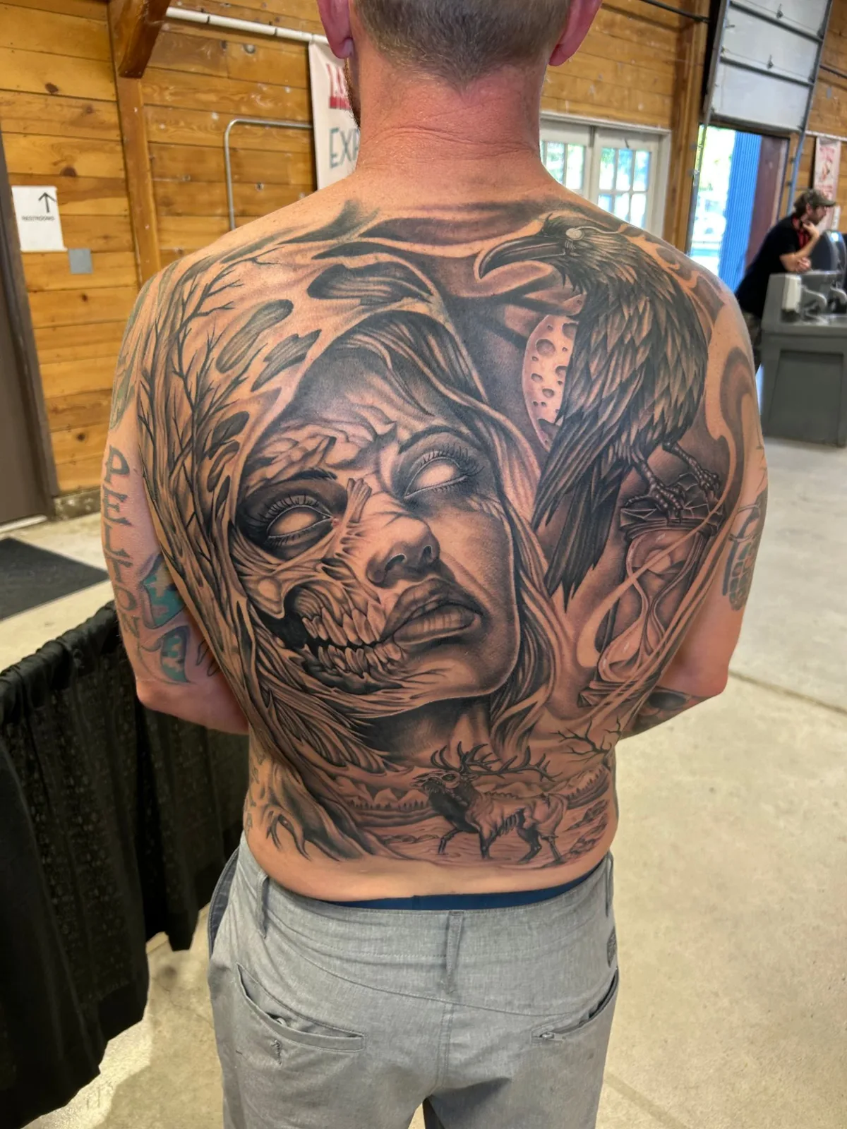Full black and grey back piece of a woman with a decaying skull face, a raven on a moon and hourglass, a bare tree and an elk by Adam Harper