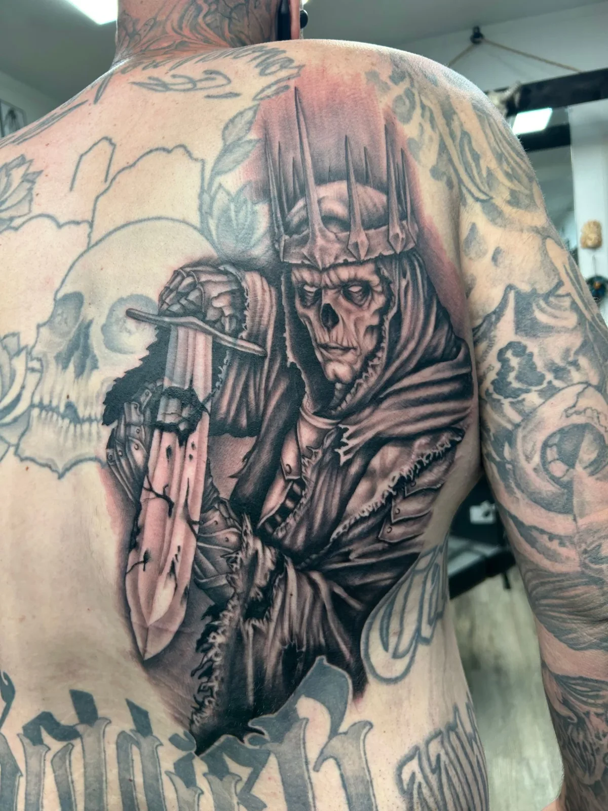 Black and grey skeletal king with a spiked crown and sword tattooed on a back by Adam Harper