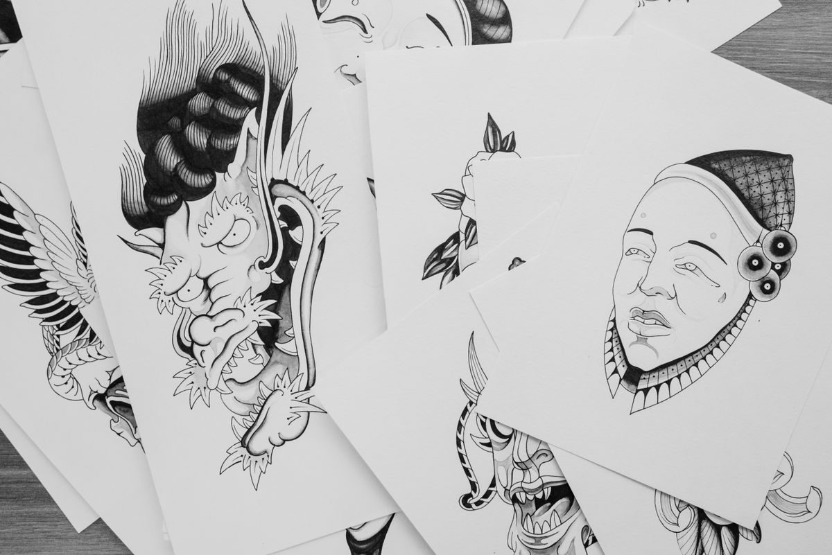 Sheets of black ink tattoo flash drawings, including a horned demon and a masked face, spread on a table