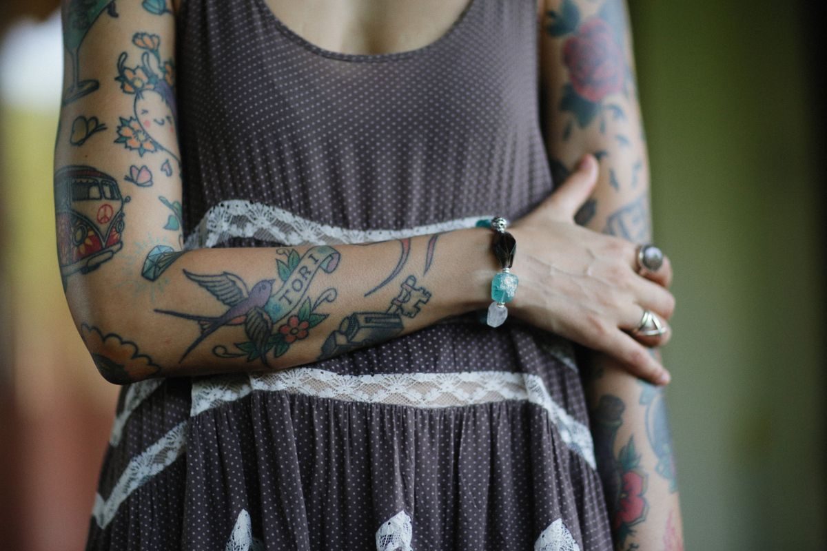 Woman in a gray dress with arms crossed, both forearms covered in tattoos