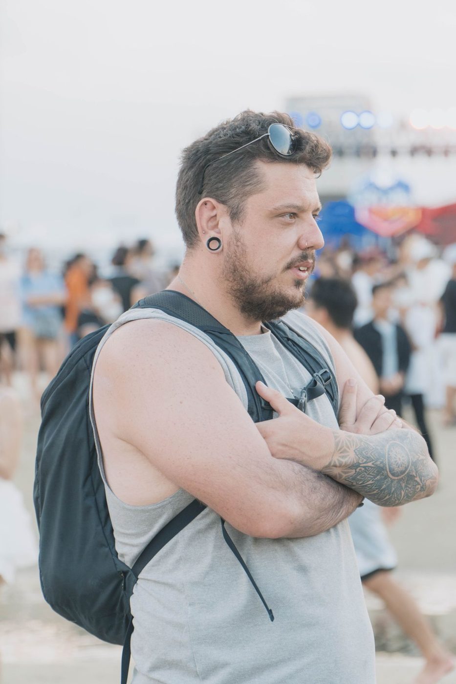 Man with arms crossed, a backpack and a tattoo on his forearm, standing at a crowded outdoor event