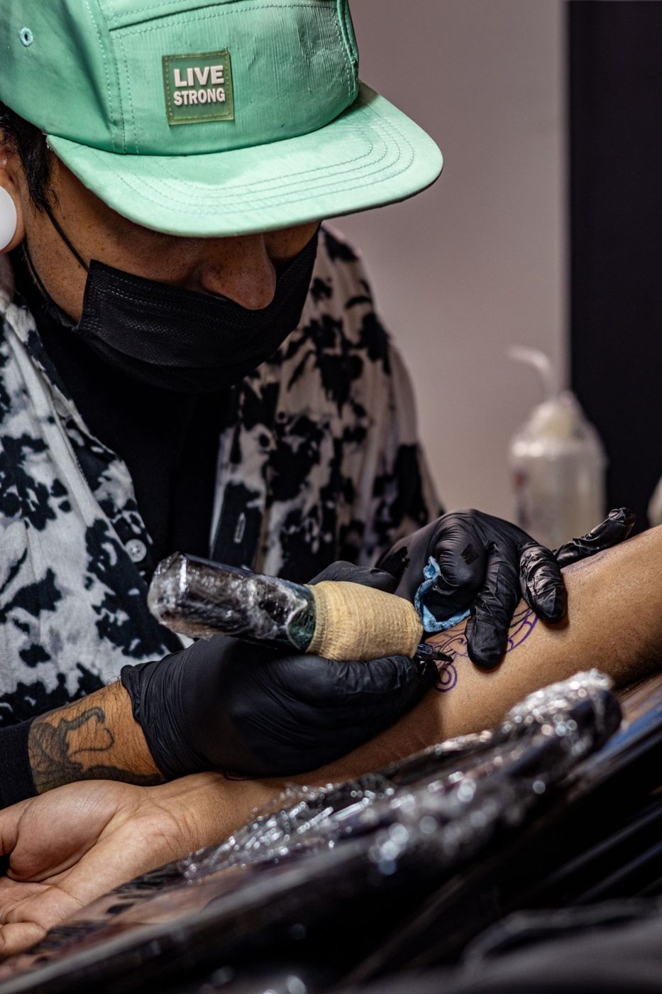 Tattoo artist in a mask and black gloves tattooing a forearm with a machine