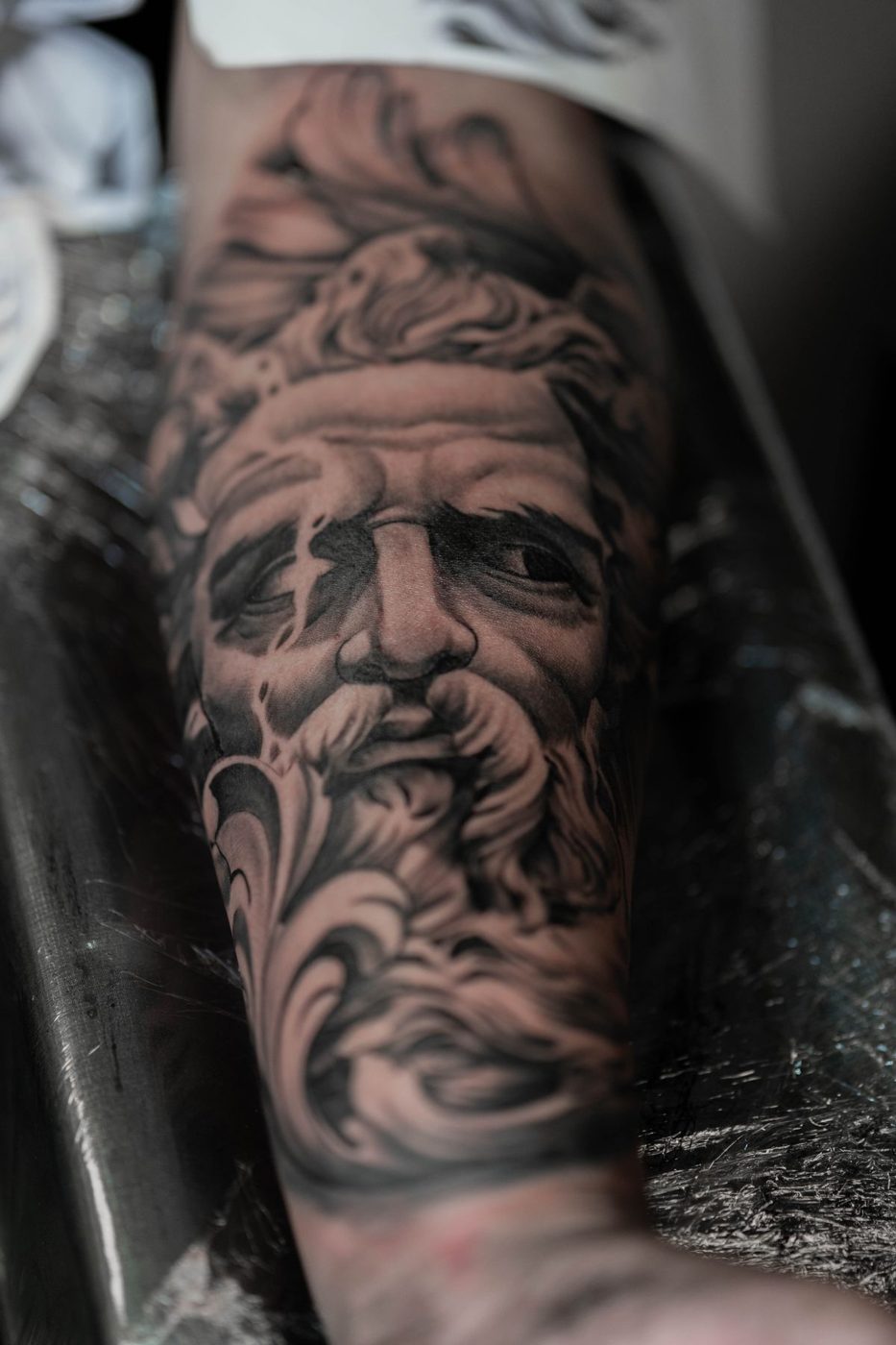 Black and grey realistic tattoo of a bearded classical-style face on a forearm
