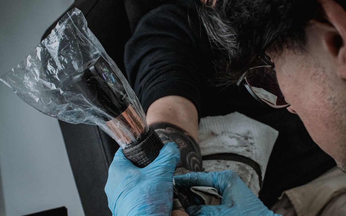 Tattoo artist in blue gloves holding clear wrap over a client's tattooed forearm