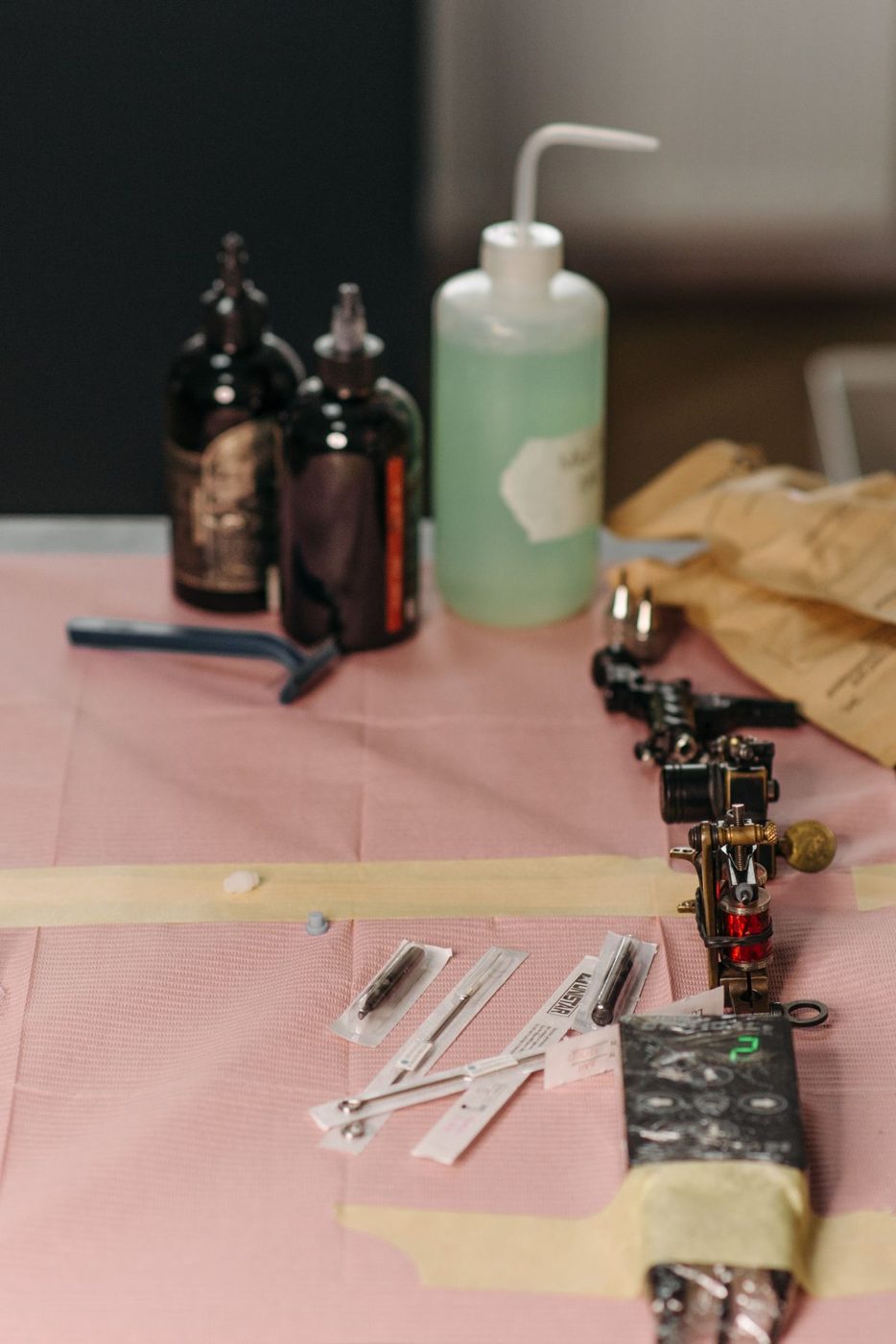 Tattoo supplies laid out on a pink barrier sheet: ink bottles, a wash bottle, sealed needle pouches and machines