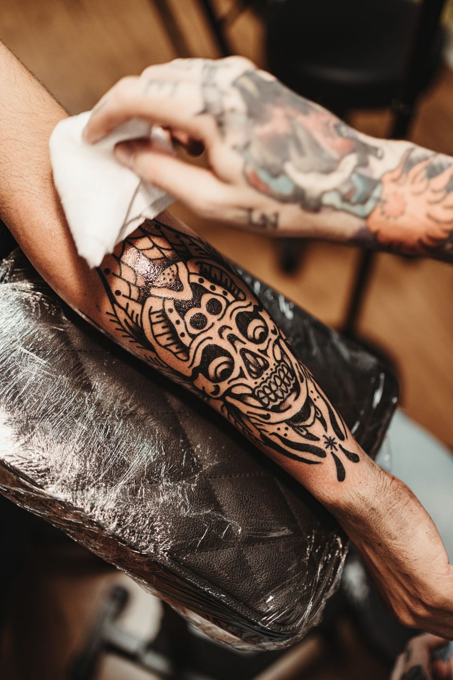 Hand pressing a paper towel against a tattooed forearm resting on a plastic-wrapped surface