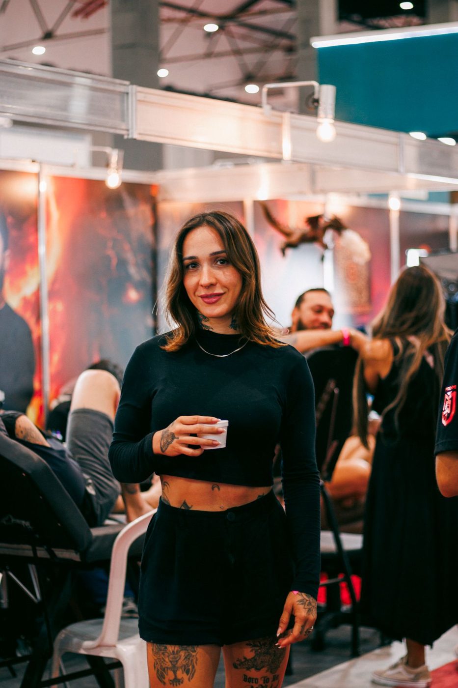 Woman holding a small cup in a tattoo convention aisle while artists work on clients behind her