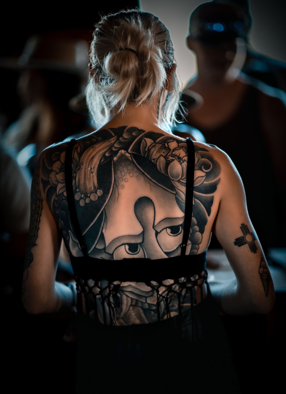 A woman with a large black and grey back tattoo seen from behind in a crowd