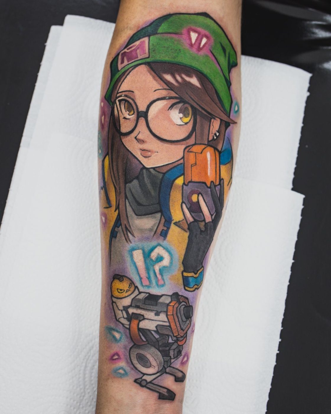 Forearm tattoo of a cartoon girl in large glasses and a green beanie in saturated colors