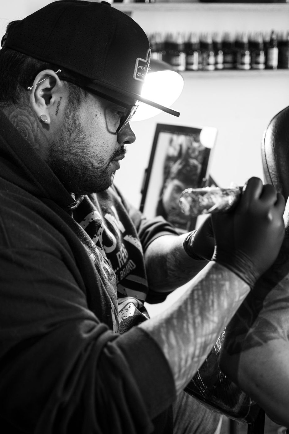 Black-and-white photo of a tattoo artist in a cap and glasses working on a client's arm
